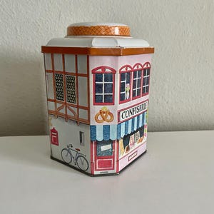 Peut inclure: Une bo&icirc;te en m&eacute;tal d&eacute;corative en forme de b&acirc;timent, avec une fa&ccedil;ade rose repr&eacute;sentant une boulangerie. La bo&icirc;te a un toit blanc et un couvercle &agrave; motifs. D&eacute;tails : un v&eacute;lo, une porte rouge et le mot "CONFIERIE".