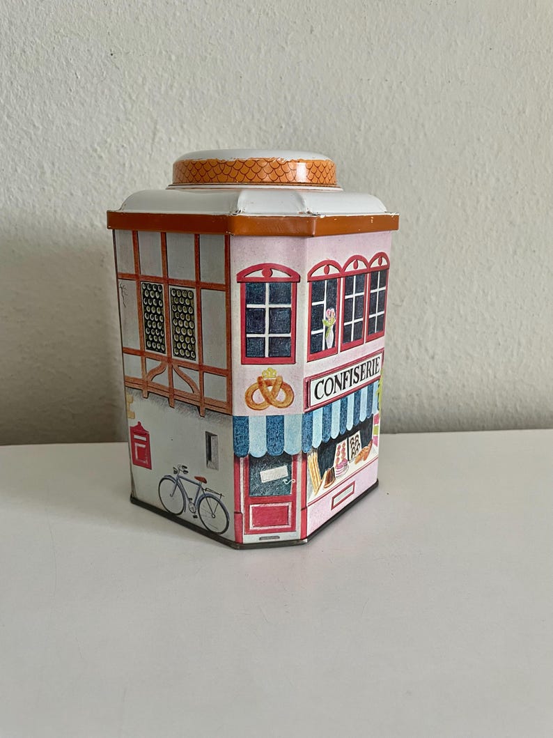 Peut inclure: Une bo&icirc;te en m&eacute;tal d&eacute;corative en forme de b&acirc;timent, avec une fa&ccedil;ade rose repr&eacute;sentant une boulangerie. La bo&icirc;te a un toit blanc et un couvercle &agrave; motifs. D&eacute;tails : un v&eacute;lo, une porte rouge et le mot "CONFIERIE".