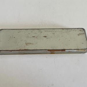 May include: A rectangular, vintage-style metal pencil case with a weathered, light grey finish. The surface shows visible signs of wear, including scratches, scuffs, and minor rust along the edges, giving it a distressed, aged appearance.