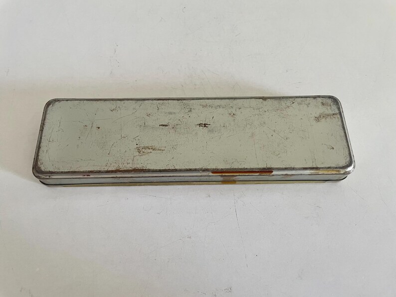 May include: A rectangular, vintage-style metal pencil case with a weathered, light grey finish. The surface shows visible signs of wear, including scratches, scuffs, and minor rust along the edges, giving it a distressed, aged appearance.