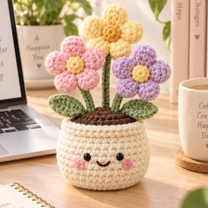 May include: A crocheted potted plant featuring three flowers in yellow, pink, and purple with green stems and leaves. The cream-colored pot has a stitched face with black eyes, a small smile, and pink cheeks, sitting on a wooden desk surface.