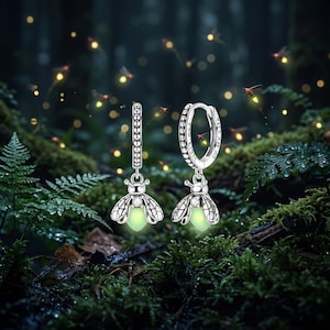 May include: A pair of silver hoop earrings featuring dangling firefly charms. Each charm has a clear stone set in the head and a glowing light green body. The hoop of one earring is engraved with the text "YOU LIGHT UP MY LIFE" in black.
