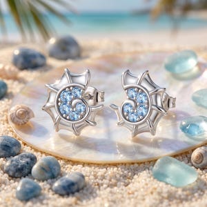 May include: A pair of silver stud earrings shaped like spiral seashells, each featuring a cluster of small, round, light blue gemstones set in the center. The earrings have a polished metallic finish and a secure post backing for attachment.