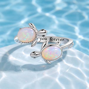 May include: A silver-toned open ring featuring two sea turtle motifs, each set with an oval iridescent opal-like stone. The band is engraved with the text you forever in a black script font, creating a continuous design across the finger.