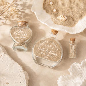 May include: Three clear glass bottles with cork stoppers filled with sand. One heart-shaped bottle features a mountain graphic and text, a round bottle displays the text HONEYMOON SAND SCOTT & OLIVIA MALDIVES 2025, and a small vial contains sand.