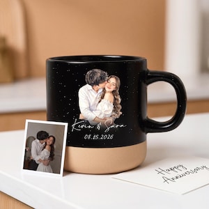 Pode incluir: Uma caneca de cerâmica preta e bege com uma ilustração personalizada de um casal a abraçar, os nomes Kevin & Sara e a data 08.15.2026 em branco. Inclui uma pequena fotografia quadrada do casal e um cartão com Happy Anniversary.