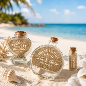 May include: Three clear glass bottles filled with sand and topped with cork stoppers. The heart-shaped bottle features a mountain graphic and the text Oliver & Sophia 06.12.2026. The round bottle reads HONEYMOON SAND Scott & Olivia MALDIVES 2025.