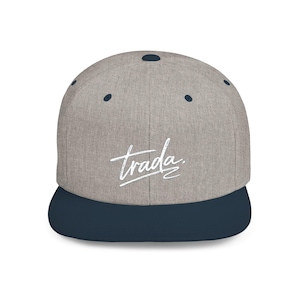 May include: A gray baseball cap with a navy blue brim. The word "trada" is written in white script across the front of the cap. The cap has a flat brim and a structured crown.