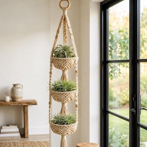 May include: A three-tiered macrame plant hanger made of off-white cord, featuring three woven baskets suspended vertically. Each basket holds a green air plant, with decorative tassels hanging between the tiers and a wooden ring at the top.