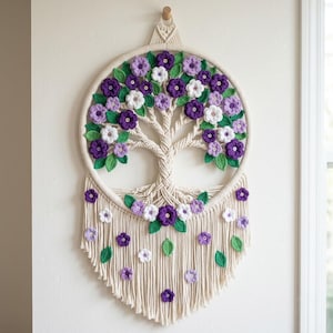 May include: A handcrafted macrame wall hanging featuring a tree design with a circular frame. The tree is adorned with purple, white, and green crocheted flowers and leaves. The piece is suspended from a wooden dowel, adding a touch of natural texture.