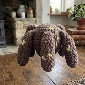 May include: A large, handmade brown crochet spider with eight thick legs and green accents. The plush toy is designed with a textured, chunky stitch and is likely intended as a decorative item or a child's toy.