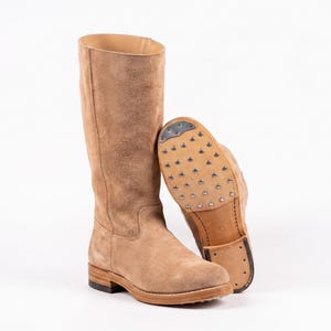 Puede incluir: Un par de botas altas de ante color beige. Una bota está de pie, mientras que la otra está tumbada de lado, mostrando la suela tachonada. Las botas tienen un diseño clásico con punta redonda y tacón apilado. Las botas miden unos 40 cm de alto.