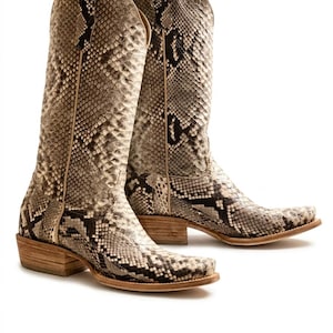 Puede incluir: Un par de botas de vaquero altas con un estampado de piel de serpiente en tonos beige, marrón y negro. Las botas tienen una puntera cuadrada, un tacón apilado y una suela de cuero. La caña de las botas es alta, llegando hasta la pantorrilla.