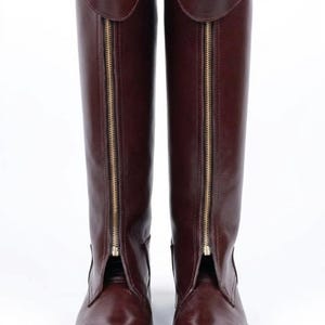 May include: A pair of tall, brown leather boots with gold zippers running down the front. The boots have a buckle detail at the top and are set against a white background. The boots are a classic equestrian style.