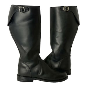 Puede incluir: Un par de botas de montar de cuero negro. Las botas son hasta la rodilla con un detalle de hebilla en la parte superior y un tacón pequeño. Las botas son de cuero liso y tienen un diseño clásico.