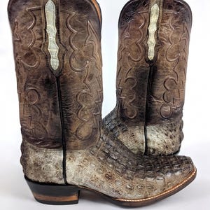 Könnte beinhalten: Ein Paar braune und beige Cowboy-Stiefel mit einem strukturierten, krokodilartigen Muster auf dem Fuß und dem unteren Bein. Der obere Schaft hat dekorative Nähte und einen hellfarbigen Akzentstreifen. Die Stiefel haben eine spitze Zehenpartie und einen gestapelten Absatz.