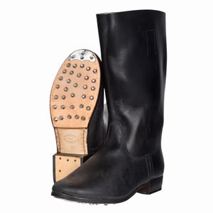 May include: Black leather knee-high boots. One boot is upright, the other is on its side, revealing the sole with metal studs. The boots have a classic design with a smooth, polished finish. The soles are made of light brown leather.