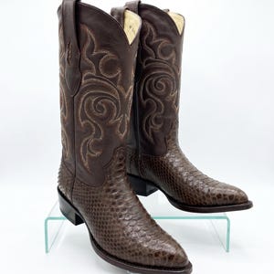 Può includere: Un paio di stivali da cowboy marroni con intricati disegni cuciti sullo stelo superiore. La parte inferiore dello stivale presenta un motivo strutturato simile alla pelle di serpente. Gli stivali sono esposti su una mensola di vetro trasparente.