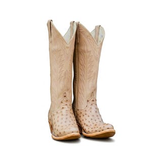 Pode incluir: Um par de botas de cowboy bege com bico fino e costuras decorativas. As botas têm um padrão texturizado no pé e uma cana alta. Os saltos têm aproximadamente 5 cm de altura.