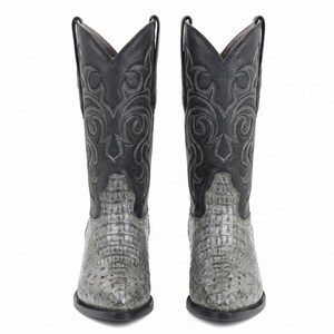 Puede incluir: Un par de botas de vaquero con un pie gris texturizado y una parte superior negra decorativa. Las botas tienen una punta puntiaguda y una caña alta, adecuadas para el uso occidental. Las botas son de cuero.