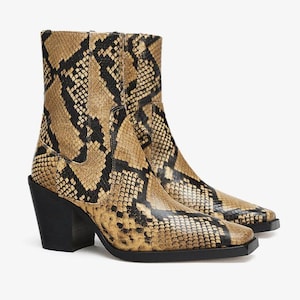 Könnte beinhalten: Stiefeletten mit einem beigen und schwarzen Schlangenledermuster. Die Stiefel haben eine quadratische Zehenpartie, einen massiven schwarzen Blockabsatz und eine schwarze Sohle. Der Schaft reicht über den Knöchel und das Äußere zeigt einen strukturierten Druck.