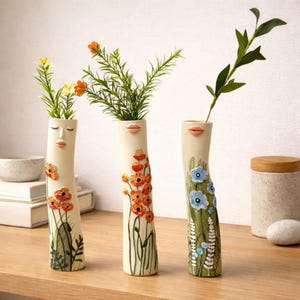 May include: Three off-white ceramic vases with painted floral designs and facial features. Each vase has a unique design, with orange, blue, and green floral accents. The vases are filled with artificial greenery and are displayed on a wooden surface.