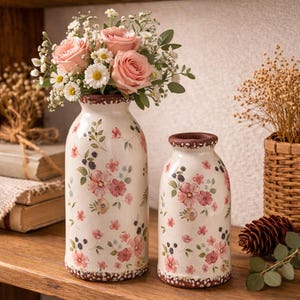 May include: Two white ceramic vases with floral designs. The larger vase holds pink roses and white daisies. The vases have a brown rim and base, and are decorated with pink flowers, green leaves, and black berries. The vases are on a wooden shelf.