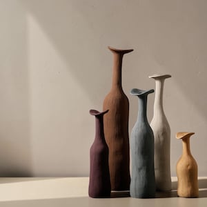 Peut inclure: Un ensemble de cinq vases hauts et élancés aux surfaces texturées et aux bords évasés et asymétriques. Les vases sont disposés à des hauteurs variables et présentent une finition mate dans des tons violet foncé, brun, bleu ardoise, blanc cassé et beige.