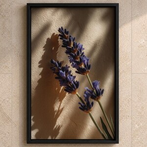 Op de afbeelding: Een ingelijste wandkunstprint met twee takjes paarse lavendelbloemen tegen een getextureerde beige achtergrond. De compositie bevat duidelijke schaduwen van de lavendelstengels en bloesems, alles gevat in een dunne zwarte lijst.