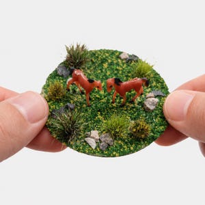 Può includere: Una base circolare per diorama in miniatura con due piccoli cavalli marroni con criniera nera su una superficie erbosa verde e strutturata. Il paesaggio include ciuffi d'erba alta, piccole pietre grigie e minuscoli accenti floreali gialli.