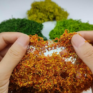 May include: A cluster of orange and yellow textured foliage material held in the foreground, with additional clumps of dark green, light green, and yellow moss-like material arranged in the background on a plain white surface.