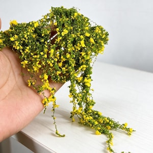 May include: A miniature trailing plant accessory featuring dense green foliage and numerous small, bright yellow flowers. The delicate, vine-like stems cascade downward, creating a realistic botanical appearance suitable for dioramas or dollhouse decor.