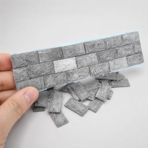 May include: A collection of small, rectangular, grey stone-textured tiles. One piece is held up to show a brick-like pattern, while several loose, individual tiles are scattered below, all featuring a rough, weathered surface finish.