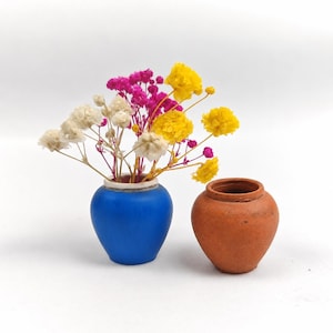 May include: A bright blue ceramic vase filled with dried white, yellow, and magenta flowers, positioned next to a small, unglazed terracotta pot with a rounded body and a wide opening, both resting on a plain white surface.