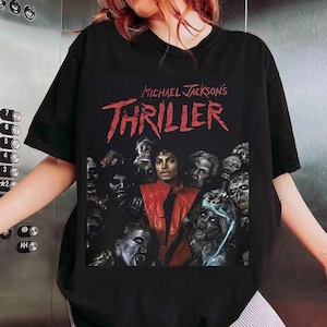 May include: Black t-shirt featuring the text "Michael Jackson's Thriller" in red, above an image of Michael Jackson in a red jacket surrounded by zombies. The shirt is a short-sleeved crew neck.