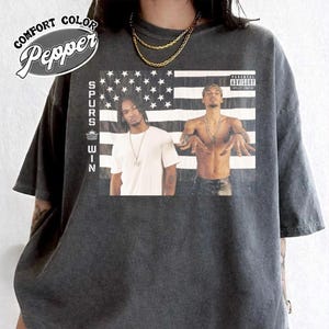 May include: Dark gray t-shirt featuring a graphic of two men in front of an American flag. The shirt has the text "SPURS WIN" vertically on the left side and "Comfort Color Pepper" in a circular design at the top left.