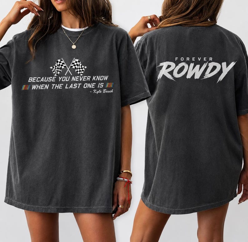 May include: Dark grey t-shirt featuring the text "BECAUSE YOU NEVER KNOW WHEN THE LAST ONE IS" and two crossed checkered flags. The back of the shirt says "FOREVER ROWDY" in white. The shirt is a casual style.
