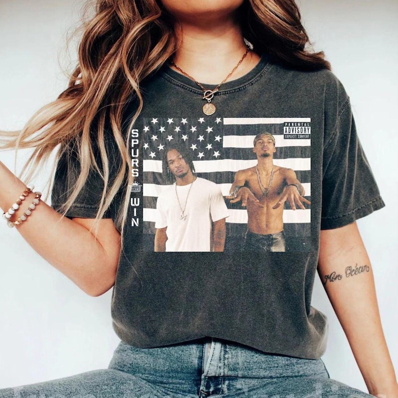 May include: Dark gray t-shirt featuring a graphic of two men in front of an American flag. The shirt has the text "SPURS WIN" on the left side and a parental advisory label. The shirt is worn by a person.