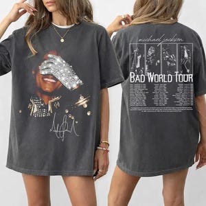 Puede incluir: Camiseta gris oscuro con un gráfico de Michael Jackson, con un guante brillante cubriendo sus ojos. La parte trasera de la camiseta muestra "BAD WORLD TOUR" y las fechas de la gira. Una camiseta de estilo vintage.