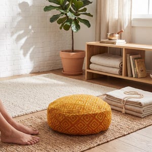 May include: A round, mustard-yellow floor cushion with a geometric diamond pattern sits on a natural fiber rug. A potted plant and a wooden shelf with folded towels and books are in the background. A yoga mat and a beige rug are also visible.