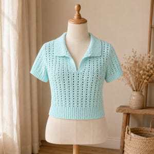 May include: A light blue, short-sleeved crochet polo shirt displayed on a mannequin. The shirt features a collared neckline and a series of vertical openwork stripes. The garment is a soft, pastel color, and the texture of the crochet is visible.