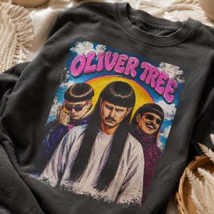 May include: A charcoal grey crew neck t-shirt featuring a graphic print of Oliver Tree. The design displays three portraits of the artist with a bowl cut hairstyle, set against a yellow sun and blue sky background with the text OLIVER TREE.