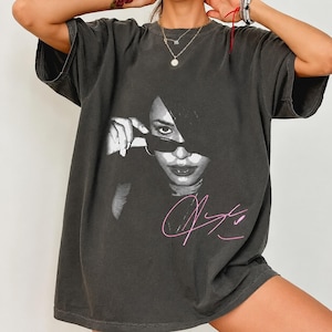May include: A charcoal gray oversized t-shirt featuring a black and white halftone graphic of a person wearing sunglasses, with a pink handwritten-style signature printed below the image. The shirt has short sleeves and a classic crew neckline.