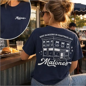 Peut inclure: T-shirt bleu marine avec le mot "Malone's" en écriture cursive sur le devant. Le dos du t-shirt présente un dessin blanc d'un bâtiment avec le texte "Best Burgers in Hastings, MA!" et le nom "Malone's".