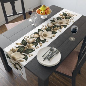 Op de afbeelding: Een witte tafelloper met een herhalende botanische print van crèmekleurige magnolia bloemen, donkergroene bladeren en goudkleurige accenten. De loper ligt op een donkere houten tafel met een stapel witte borden en wijnglazen.