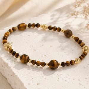 Pode incluir: Uma pulseira de contas com pedras de olho de tigre e detalhes dourados. A pulseira tem uma mistura de contas redondas pequenas e grandes, com contas espaçadoras douradas em forma de cubo e redondas. A pulseira é exibida em uma superfície branca texturizada.