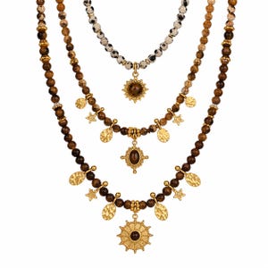May include: Three layered necklaces with beaded strands in brown and cream tones. The necklaces feature gold-colored pendants, including sunbursts, oval shapes, and star and disc charms. The beads vary in size and color, creating a layered effect.