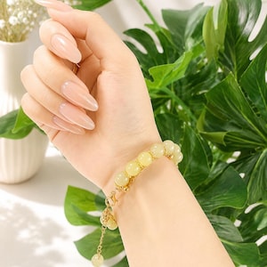 Op de afbeelding: Een goudkleurige armband met gele kralen wordt om een pols gedragen. De armband heeft een reeks ronde, lichtgele kralen die met goudkleurige schakels zijn verbonden. Een bedel hangt aan de sluiting. De achtergrond omvat groene bladeren en witte bloemen.