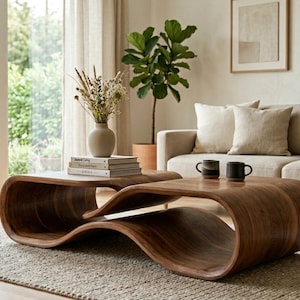 May include: A modern living room scene featuring two organically shaped wooden coffee tables. The tables have a rich, dark wood finish and sit on a neutral-toned rug. A beige sofa with pillows and a blanket is in the background, along with a potted plant and a window.