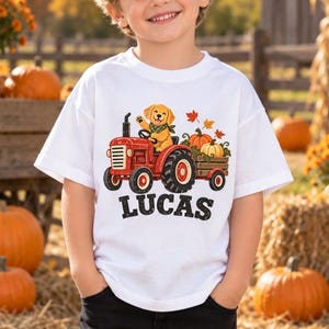 May include: A white short-sleeve t-shirt featuring a graphic of a golden retriever puppy wearing a green bandana, driving a red tractor that is pulling a wooden trailer filled with orange pumpkins and autumn leaves, with the name LUCAS printed below.